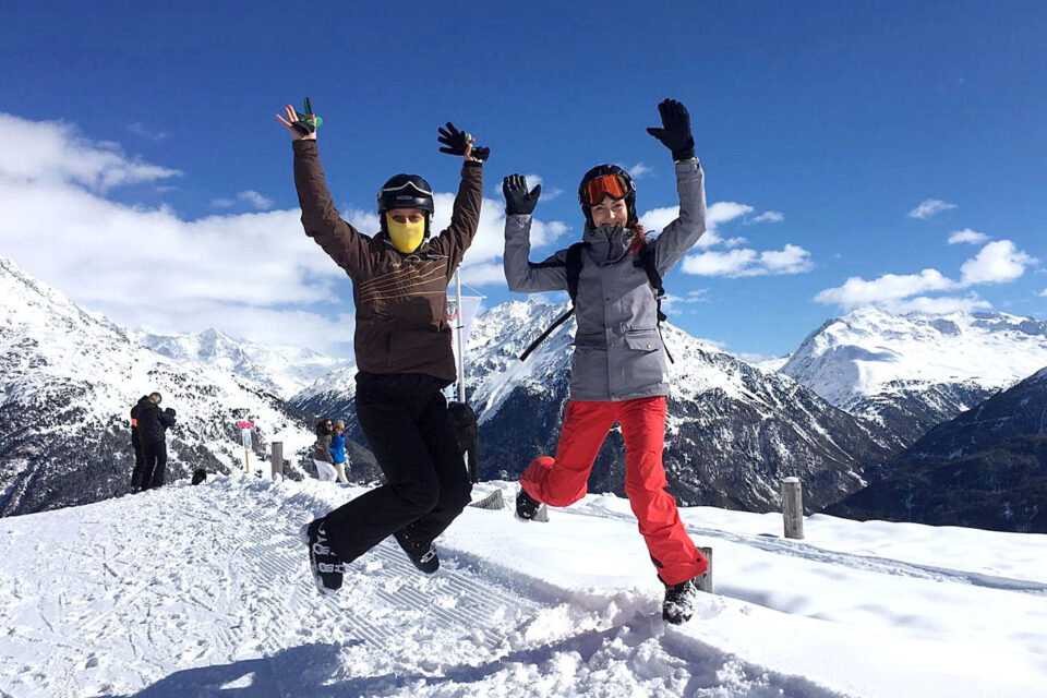 Zwei Menschen mit Skibrillen und Winterkleidung springen lachend auf einer verschneiten Berglandschaft in die Luft.