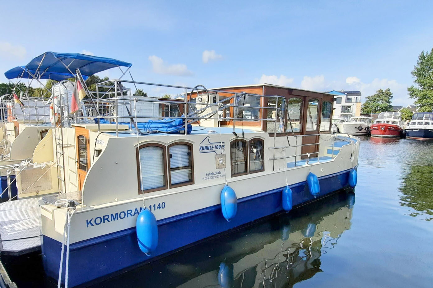 Ein weiß-blaues Boot mit Holzaufbau liegt an einem ruhigen Hafenbecken unter klarem Himmel.