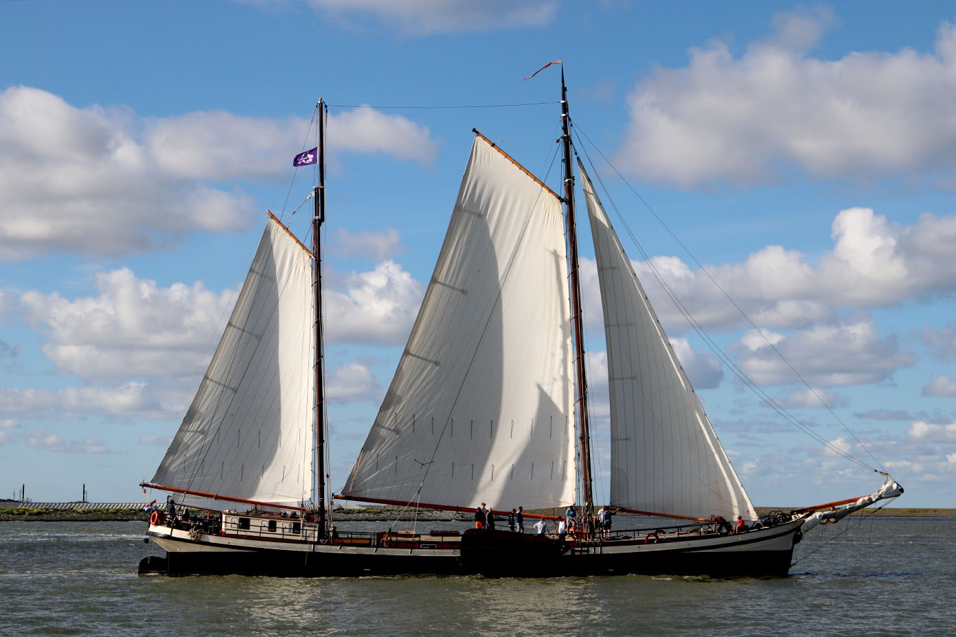 Ein Segelschiff mit drei großen weißen Segeln fährt auf ruhigem Wasser unter einem blauen Himmel mit vereinzelten Wolken.