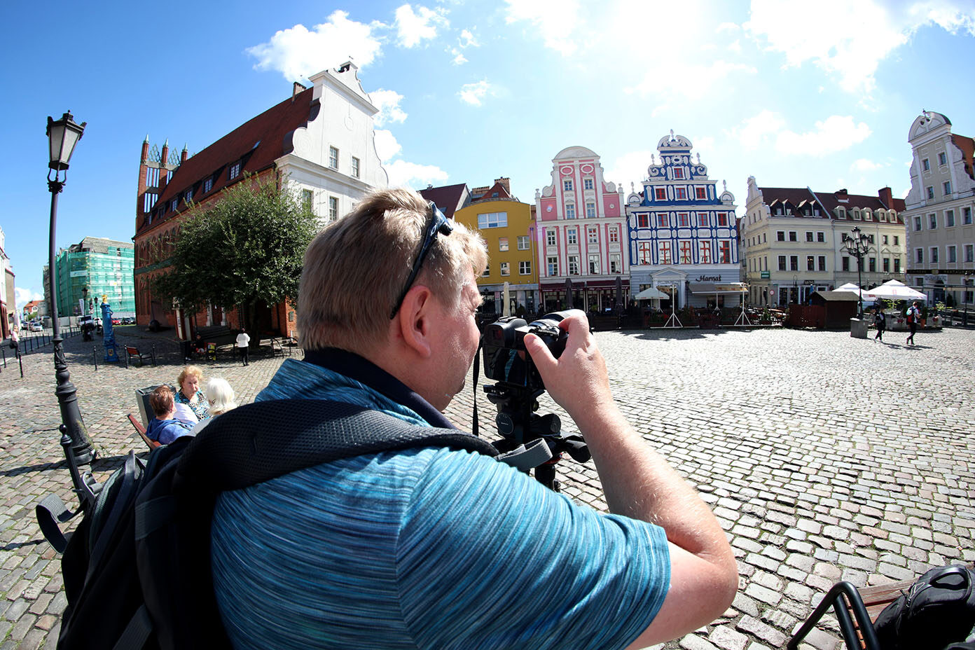 Eine Person mit blauem gestreiftem Shirt und Rucksack fotografiert auf einem gepflasterten Platz bunte historische Gebäude bei sonnigem Himmel.
