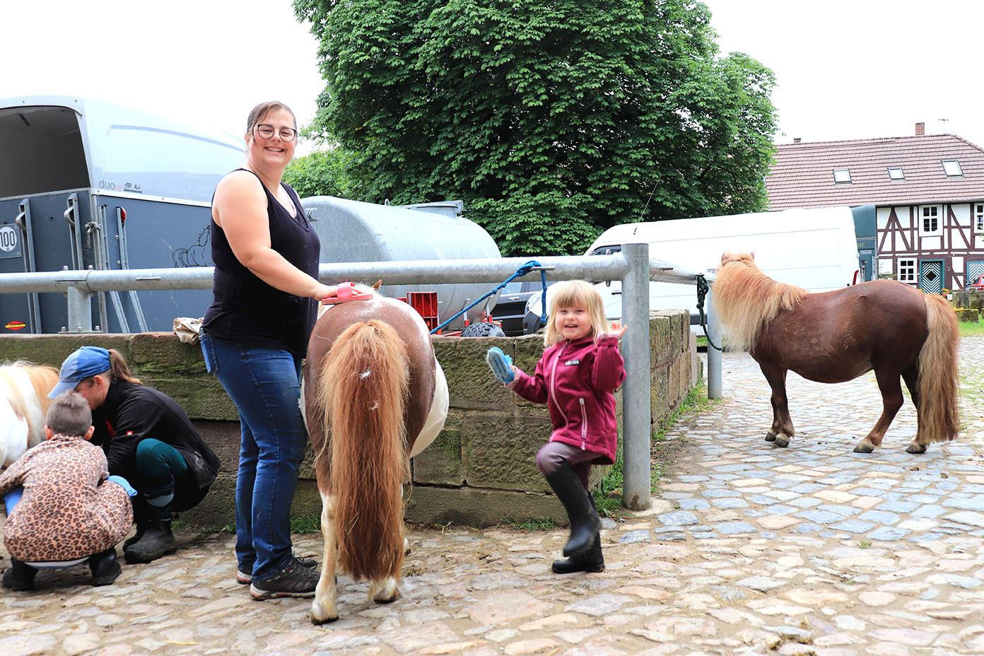 Mehrere Personen, darunter ein Kind mit Gummistiefeln und eine Person mit Brille, bürsten zwei Ponys auf einem gepflasterten Hof mit Gebäuden und Fahrzeugen im Hintergrund.