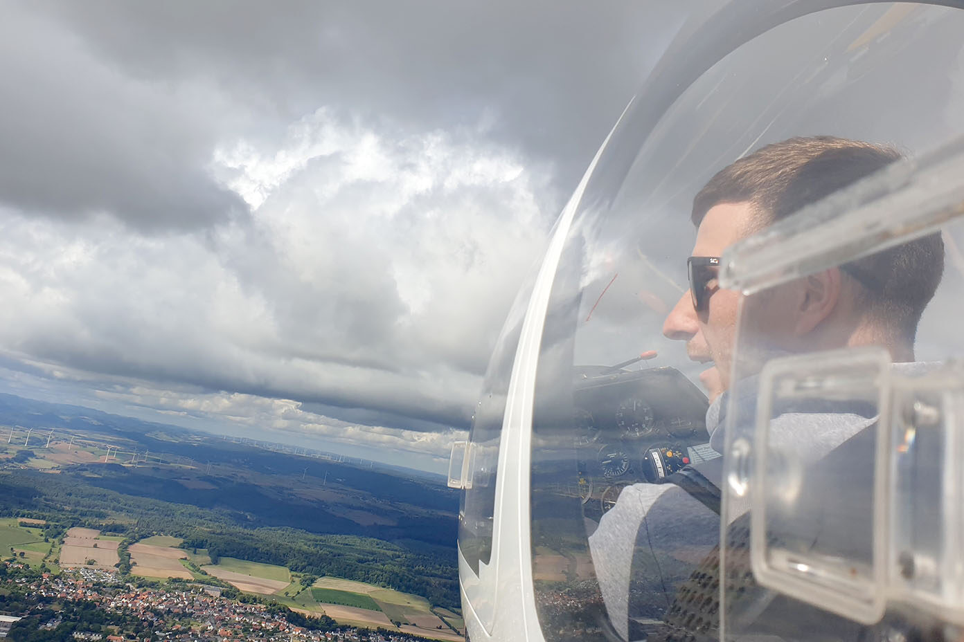 Eine Person mit Sonnenbrille sitzt in einem Segelflugzeug und blickt durch die transparente Haube auf eine Landschaft mit Feldern und Wolken am Himmel.