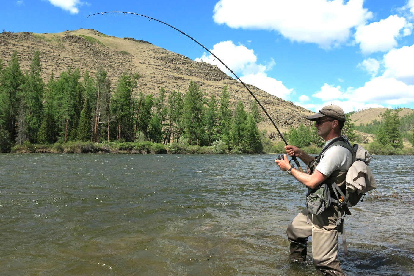 Eine Person mit Outdoor-Kleidung und Rucksack steht im Fluss und angelt mit einer gebogenen Angelrute vor bewaldeten Hügeln unter blauem Himmel mit Wolken.