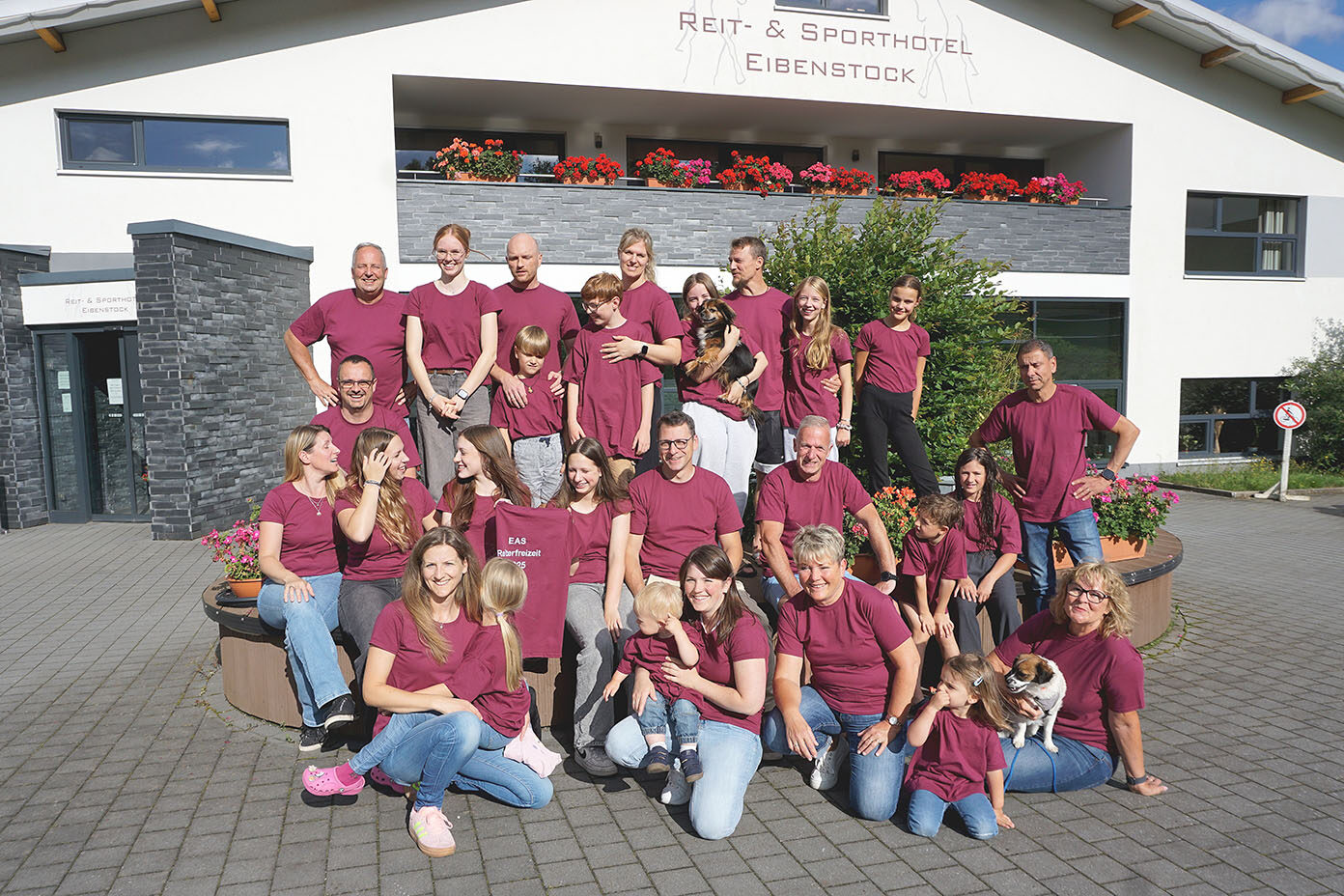 Eine Gruppe von etwa 25 Personen in einheitlichen dunkelroten T-Shirts posiert fröhlich vor einem Hotelgebäude mit Blumen und einem kleinen Hund.
