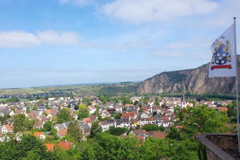 Eine malerische Stadt mit vielen Häusern vor einem bewaldeten Hügel und einem Felsen im Hintergrund unter einem blauen Himmel mit vereinzelten Wolken.