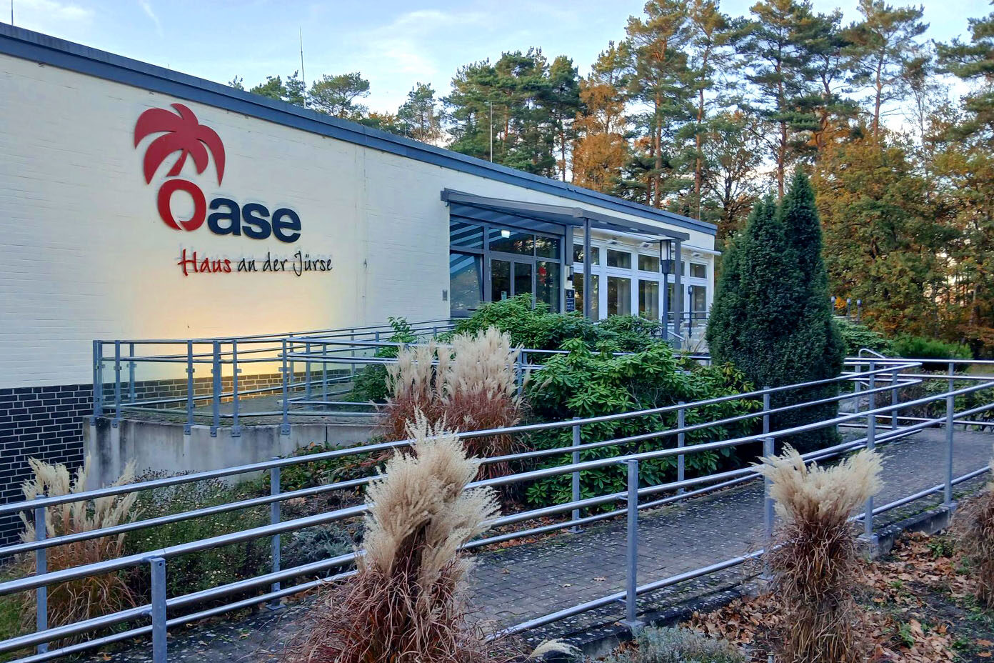 Ein Gebäudeeingang mit dem Schriftzug "Oase Haus an der Jürse" ist von einem Garten mit Büschen und einem Weg mit Geländer umgeben, im Hintergrund sind Bäume zu sehen.
