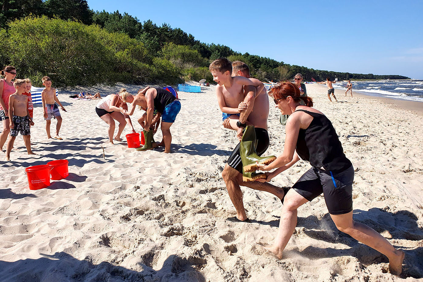 Mehrere Personen spielen am Strand ein Spiel mit roten Eimern und Sand bei sonnigem Wetter.