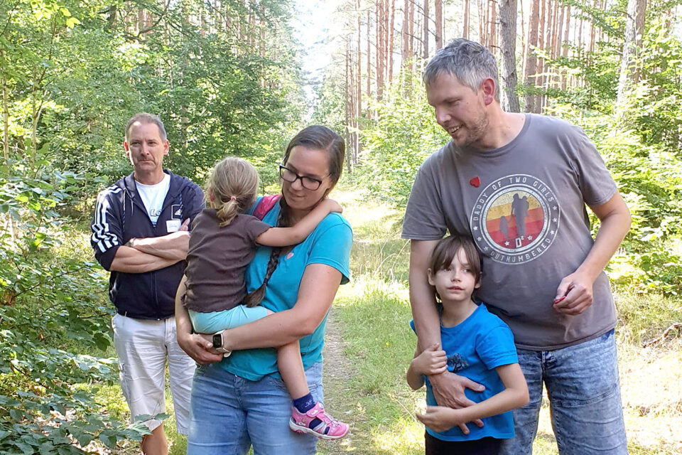 Fünf Personen, darunter zwei Kinder, stehen in einem sonnigen Waldweg, wobei eine Person ein Kind im Arm hält und eine andere Person ein Kind umarmt.