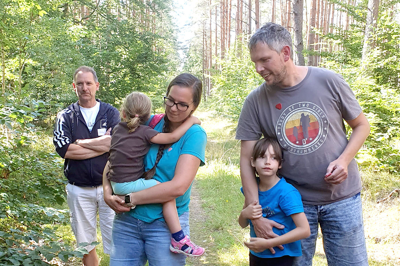 Fünf Personen, darunter zwei Kinder, stehen in einem sonnigen Waldweg, wobei eine Person ein Kind im Arm hält und eine andere Person ein Kind umarmt.