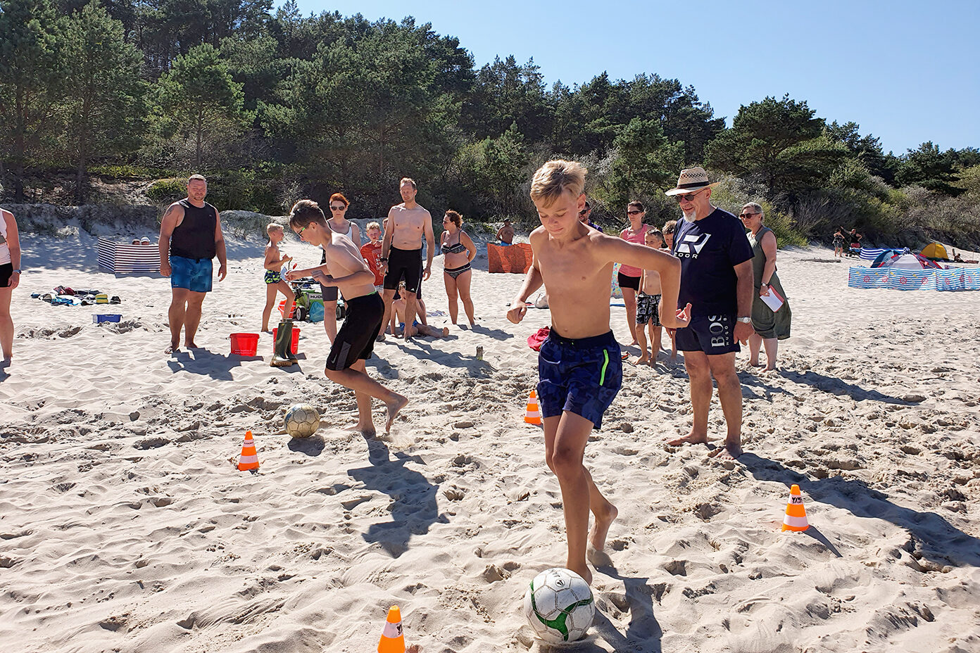 Junge im Badeoutfit dribbelt einen Fußball durch Hütchen auf einem Sandstrand, umgeben von mehreren Menschen im Hintergrund.