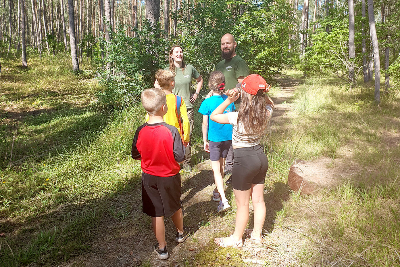 Mehrere Kinder stehen auf einem Waldweg und hören zwei Erwachsenen zu, die ihnen etwas erklären.