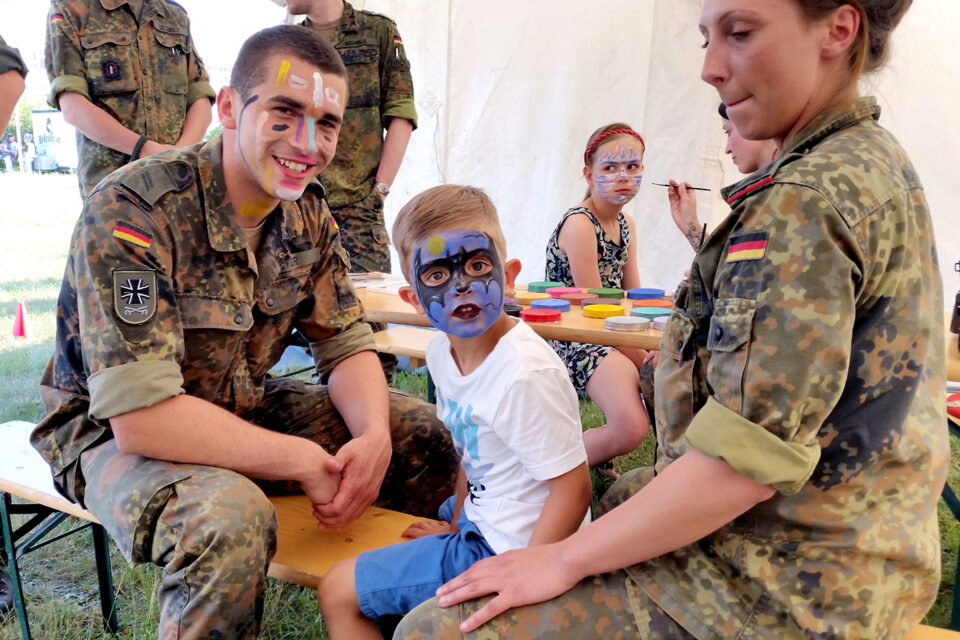 Mehrere Personen in Tarnuniform sitzen und unterhalten sich mit Kindern, die geschminkt werden, die an einem Tisch mit bunten Farben sitzen, in einem Zelt bei Tageslicht.