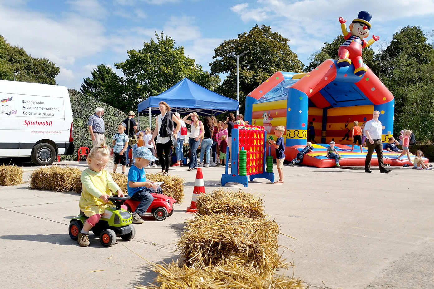 Kinder fahren auf kleinen Tretfahrzeugen auf einem mit Strohballen abgesteckten Platz vor einer bunten Hüpfburg mit Clownfigur und mehreren Erwachsenen und Kindern im Hintergrund