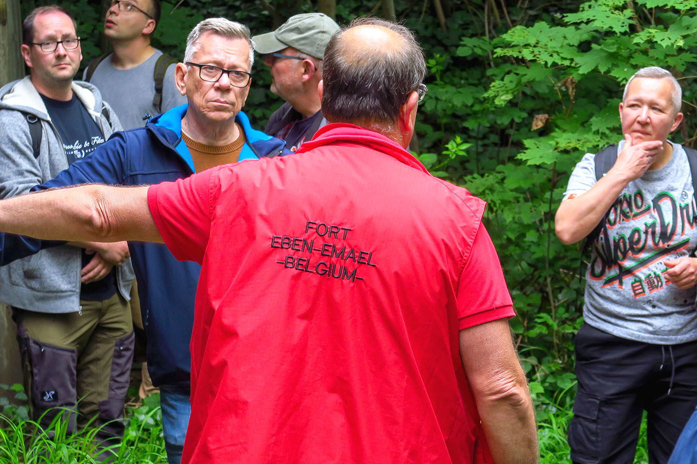 Eine Person in roter Weste mit der Aufschrift Fort Eben-Emael Belgium spricht zu einer Gruppe von Menschen vor einem grünen Waldhintergrund.