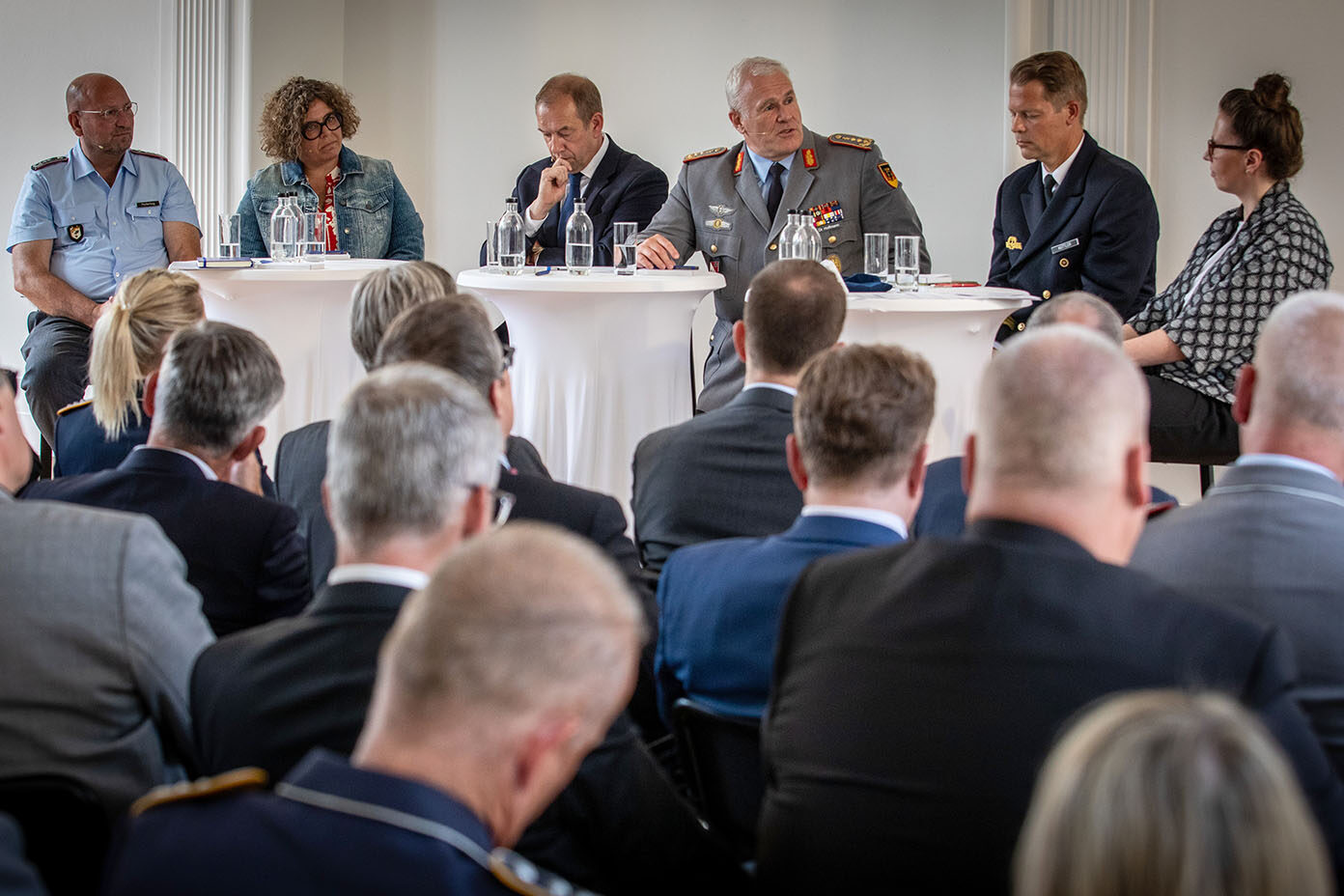 Sechs Personen sitzen an runden Tischen und diskutieren vor einem Publikum in einem Konferenzraum.