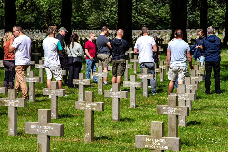 Mehrere Erwachsene stehen zwischen Reihen von Grabkreuzen auf einem Soldatenfriedhof, umgeben von Bäumen und Gras.