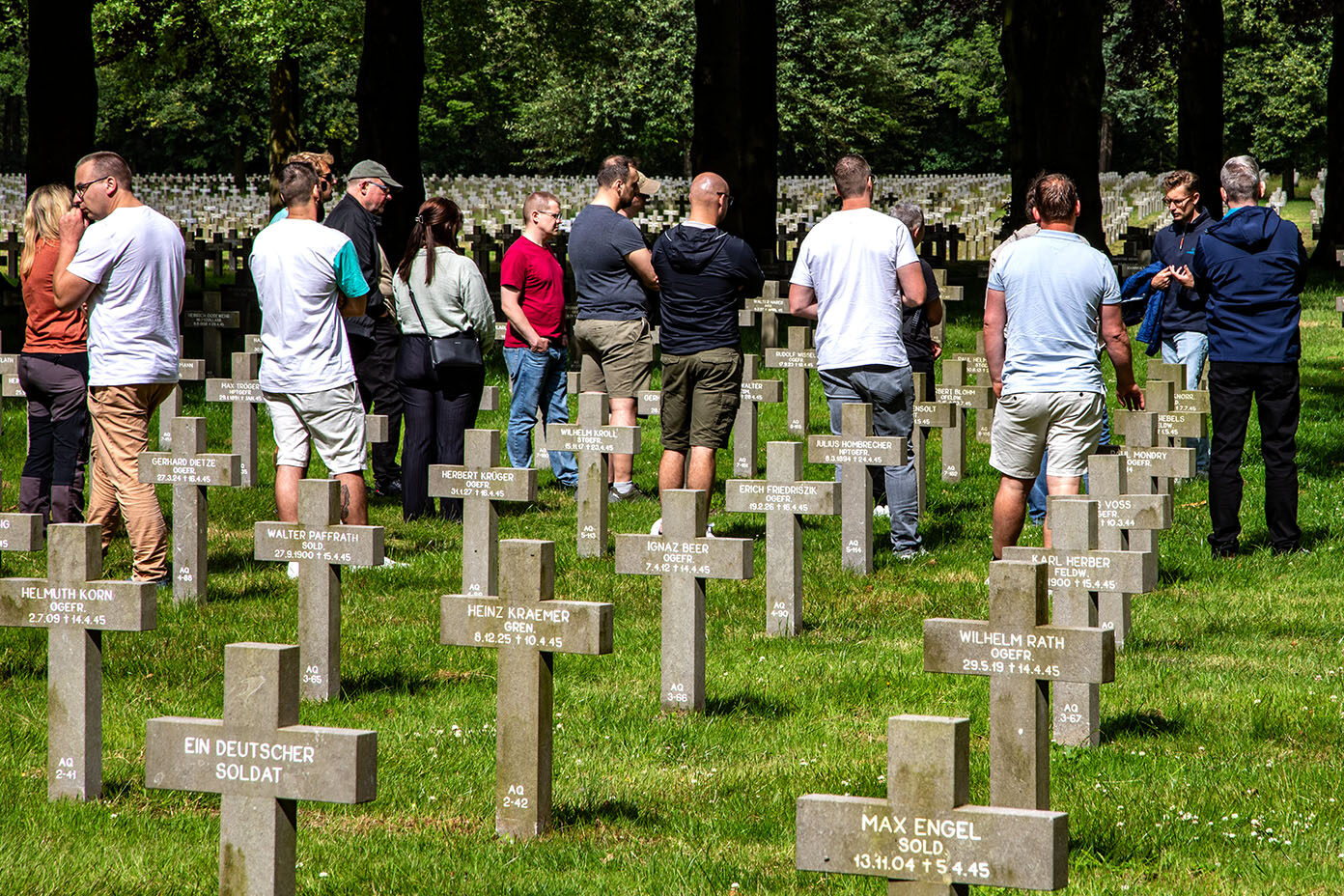 Mehrere Erwachsene stehen zwischen Reihen von Grabkreuzen auf einem Soldatenfriedhof, umgeben von Bäumen und Gras.