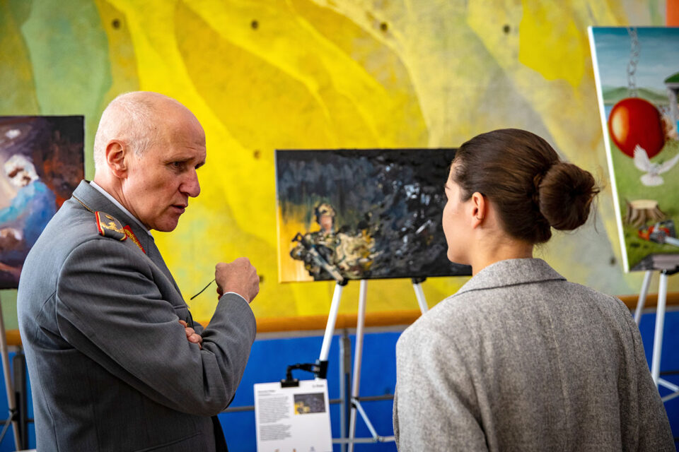 Zwei Personen, ein General und eine Zivilperson, betrachten und diskutieren vor Gemälden in einer Ausstellung.