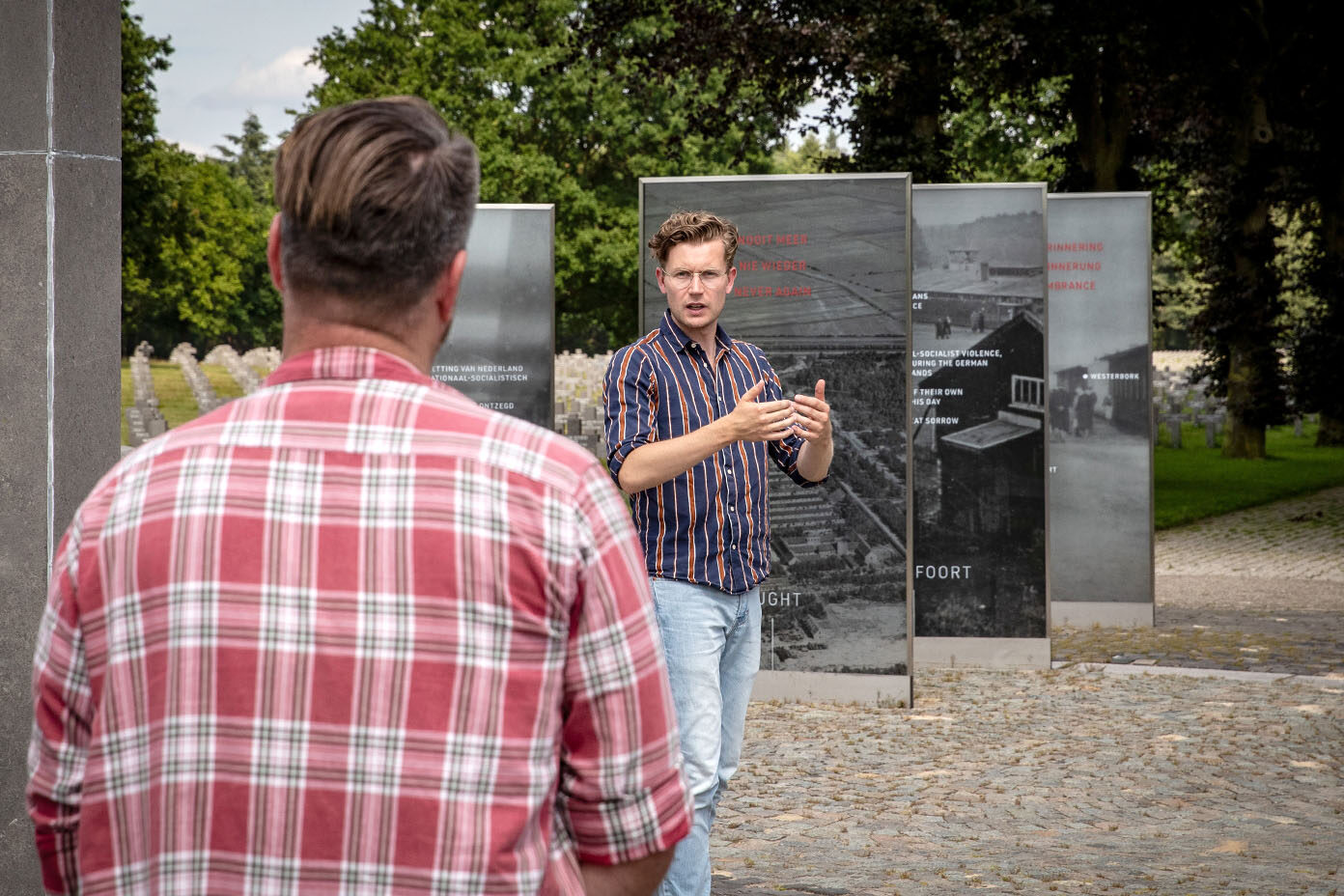 Ein Mann in gestreiftem Hemd spricht gestikulierend vor mehreren Informationstafeln im Freien, während eine weitere Person im karierten Hemd im Vordergrund steht.