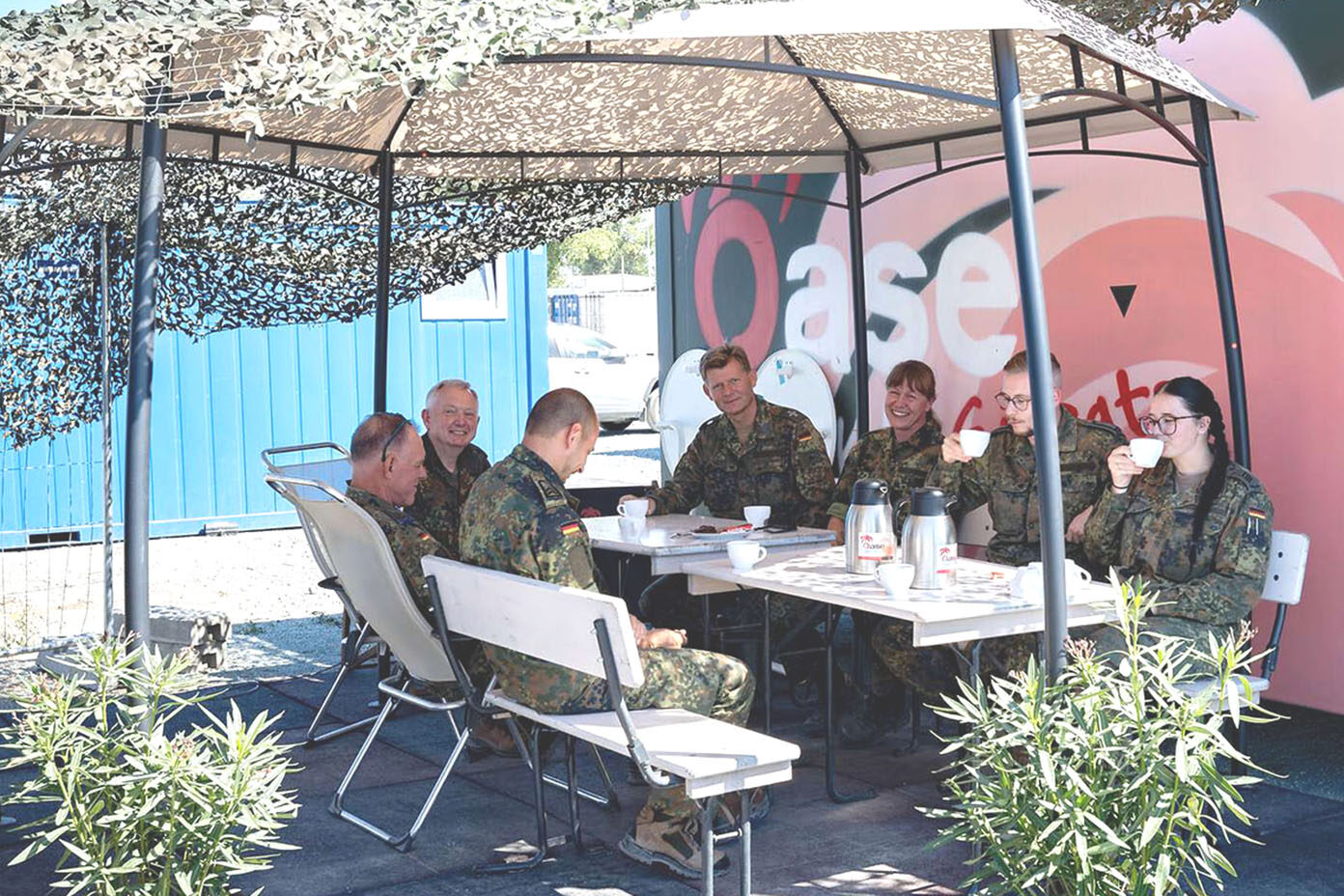 Mehrere Personen in Tarnuniformen sitzen an einem Tisch unter einem Sonnenschutz aus Tarnnetz.