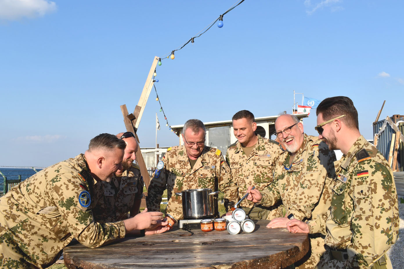 Sechs Soldaten in Tarnuniformen stehen um einen runden Holztisch im Freien und lachen, auf dem ein großer Kochtopf und mehrere Dosen stehen.