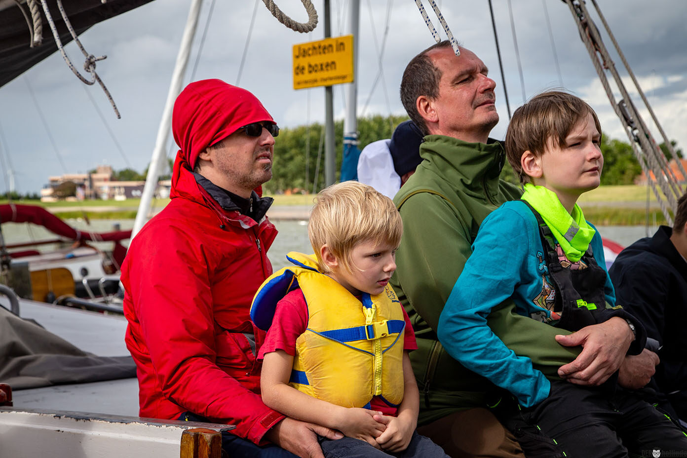 Mehrere Personen in bunter Outdoor-Bekleidung sitzen auf einem Segelboot, wobei ein Kind eine gelbe Schwimmweste trägt.