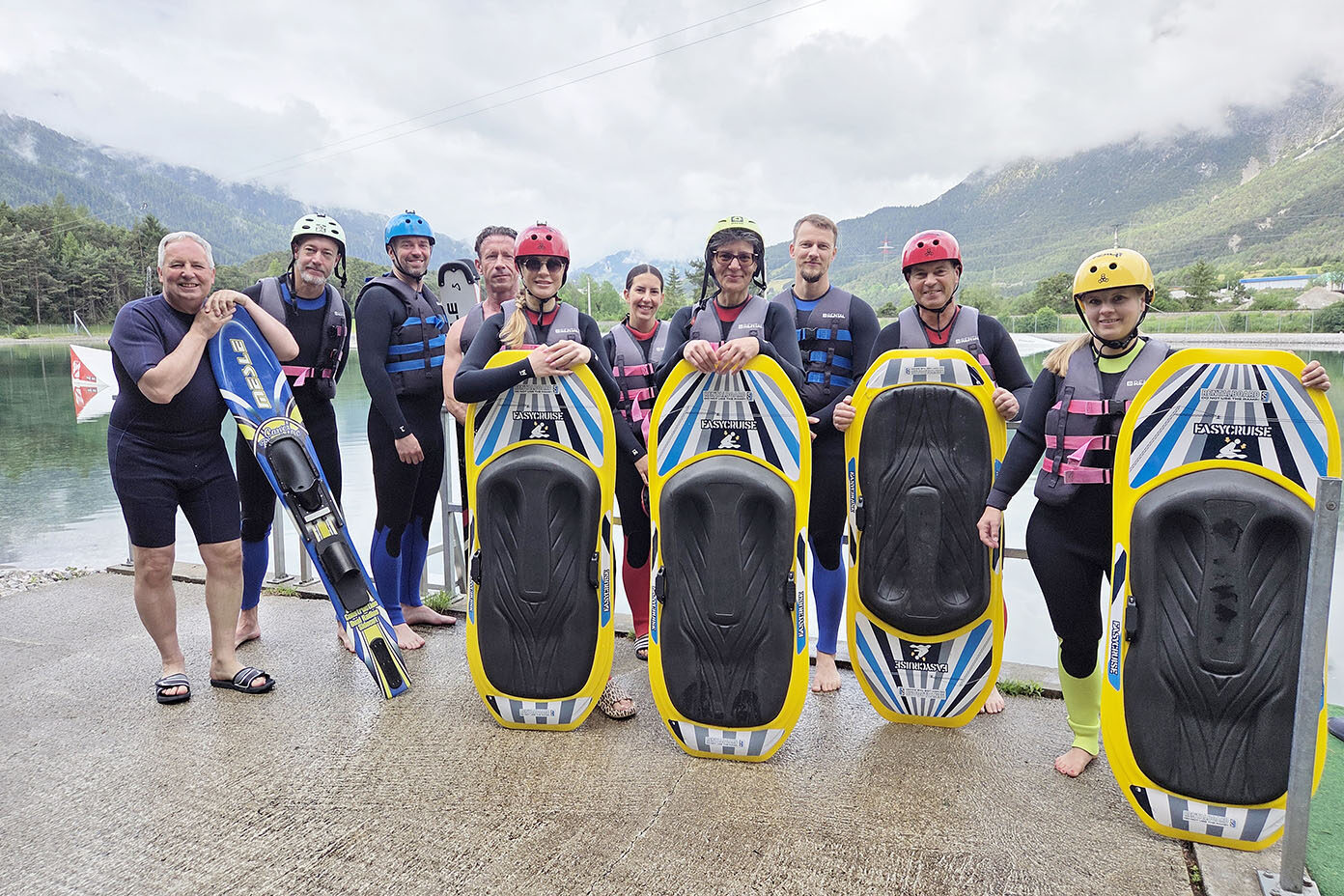 Eine Gruppe von Menschen in Neoprenanzügen und Helmen steht am Ufer eines Sees vor einer Berglandschaft und hält Wassersportgeräte.