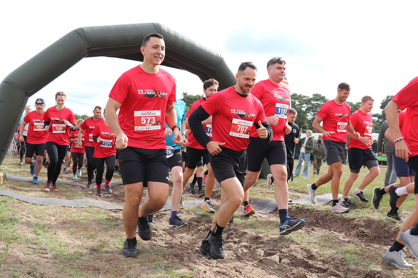 Mehrere Erwachsene in roten T-Shirts laufen bei einem Outdoor-Laufwettbewerb auf einem unebenen, schlammigen Weg unter einem aufblasbaren Startbogen.