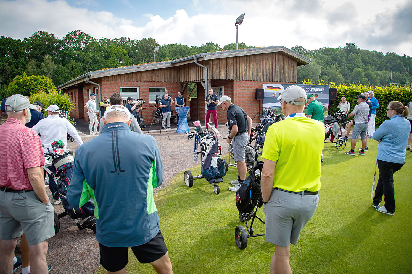 Mehrere Personen in sportlicher Kleidung versammeln sich mit Golfausrüstung vor einem kleinen Holzgebäude auf einem Golfplatz an einem bewölkten Tag.