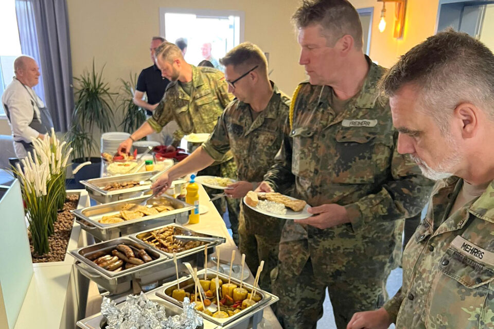 Mehrere Personen in Militäruniformen stehen an einem Buffet mit verschiedenen warmen Speisen und bedienen sich mit ihren Tellern am Essen.