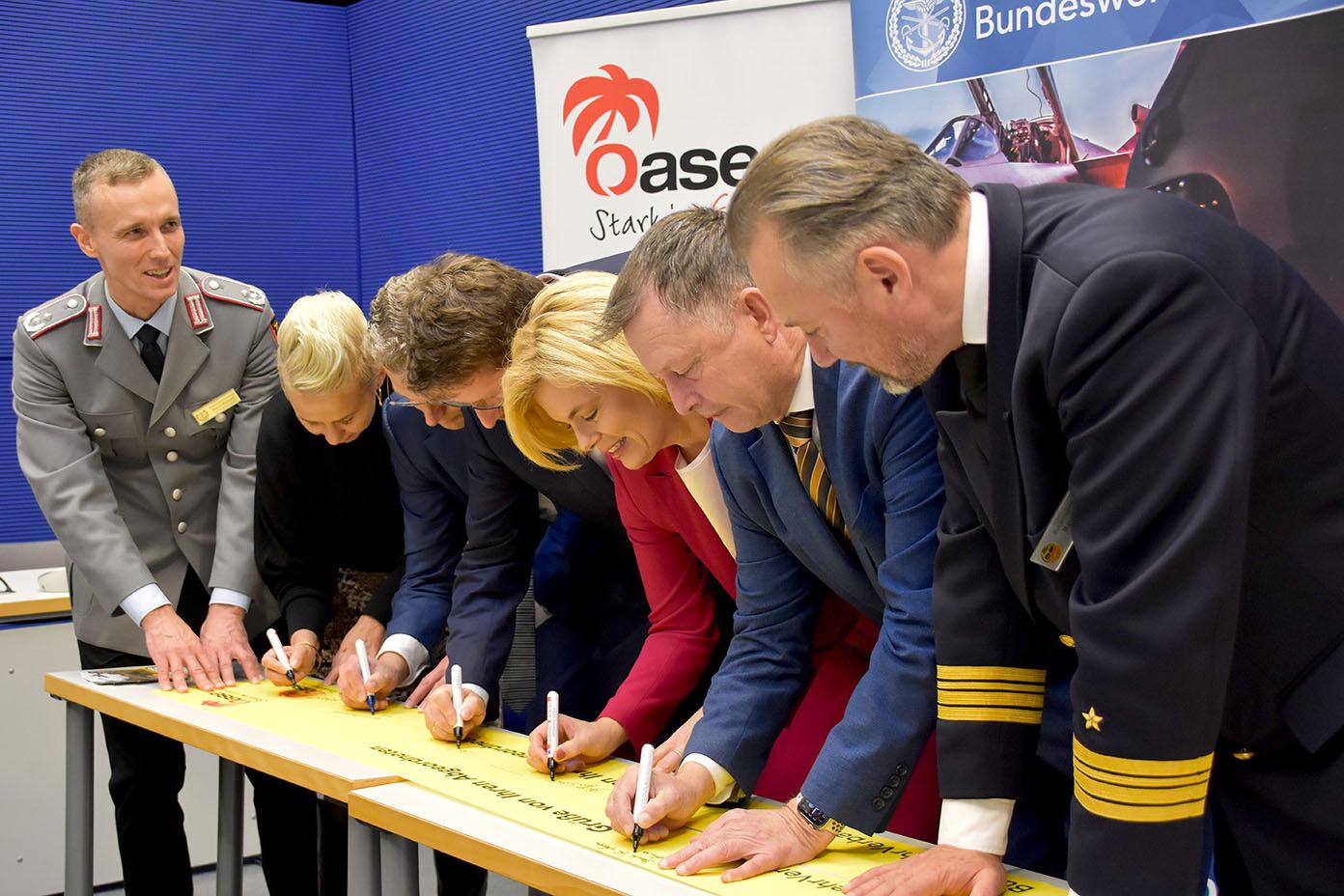Mehrere Abgeordnete des Deutschen Bundestages unterschreiben gemeinsam ein langes gelbes Band auf einem Tisch, das von zwei Personen in Uniform festgehalten wird.