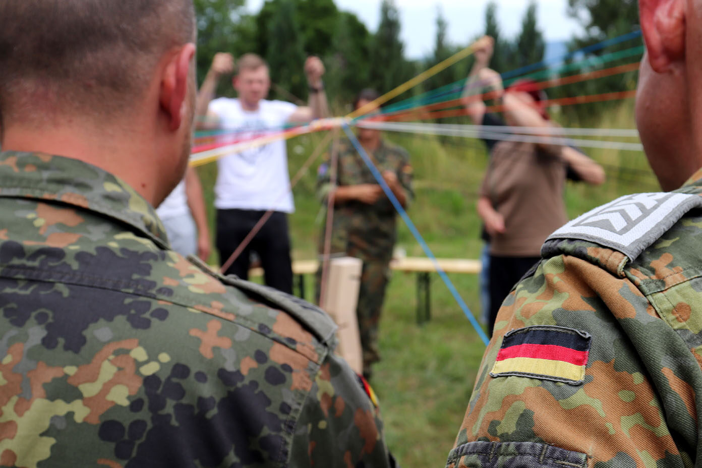 Zwei Soldaten in Tarnuniform mit deutschen Flaggen auf den Ärmeln stehen im Vordergrund, im Hintergrund mehrere Personen, die bunte Schnüre halten