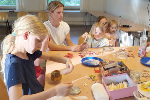 Zwei Erwachsene und drei Kunder sitzen an einem Tisch und basteln mit verschiedenen Materialien wie Papier, Pinseln, Hölzern und Muscheln in einem hellen Raum mit Fenstern.