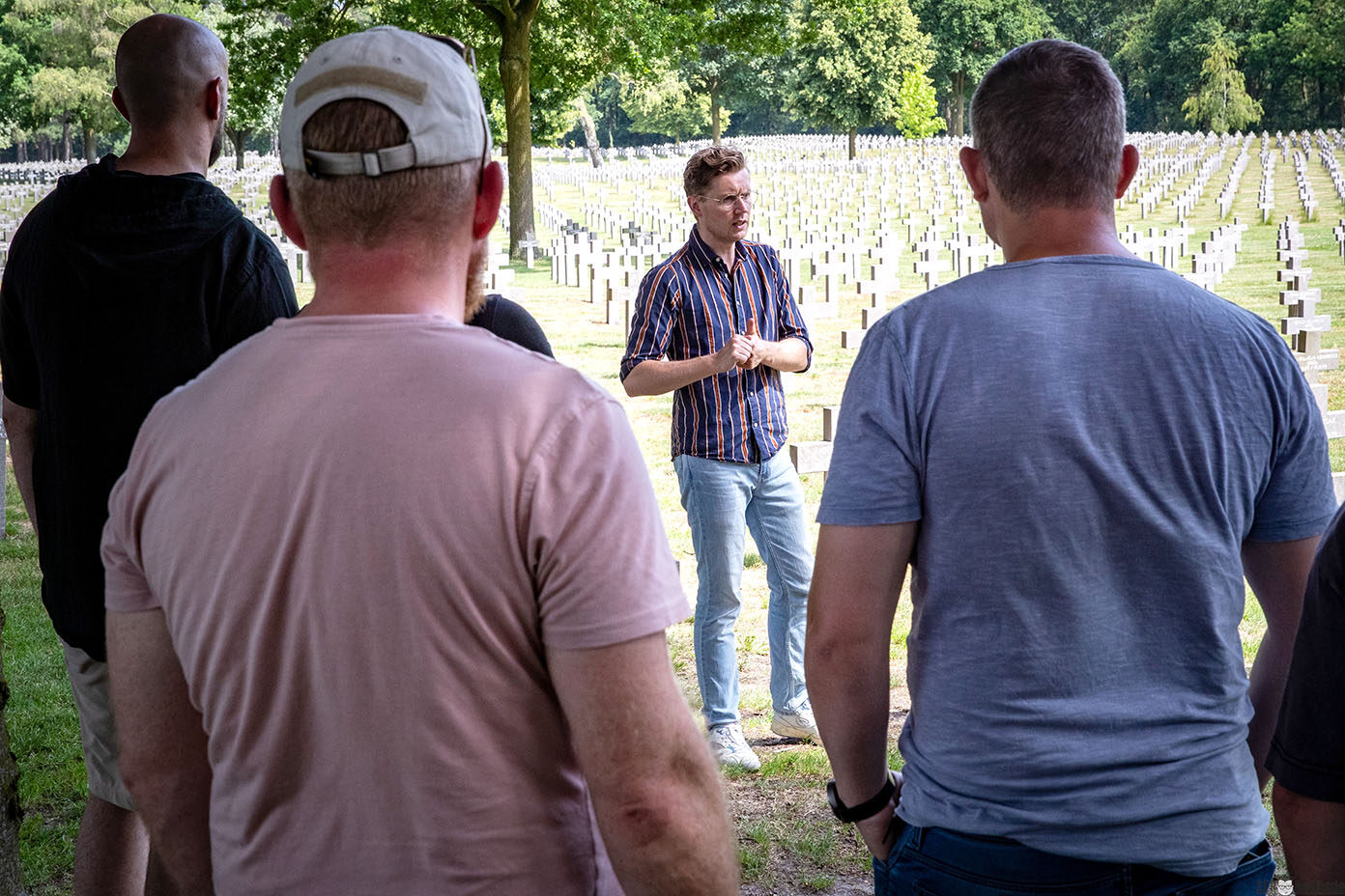 Mehrere Personen stehen auf einem Soldatenfriedhof mit vielen Grabkreuzen, während eine Person in einem gestreiften Hemd vor ihnen spricht.