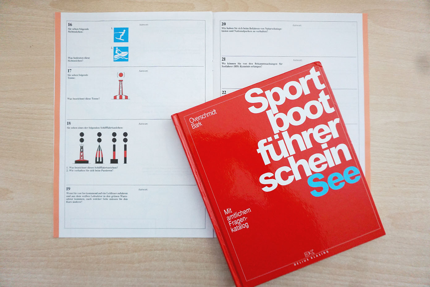 Ein rotes Buch mit dem Titel Sportbootführerschein See liegt auf einem geöffneten Lehrbuch mit nautischen Symbolen auf einem Tisch.