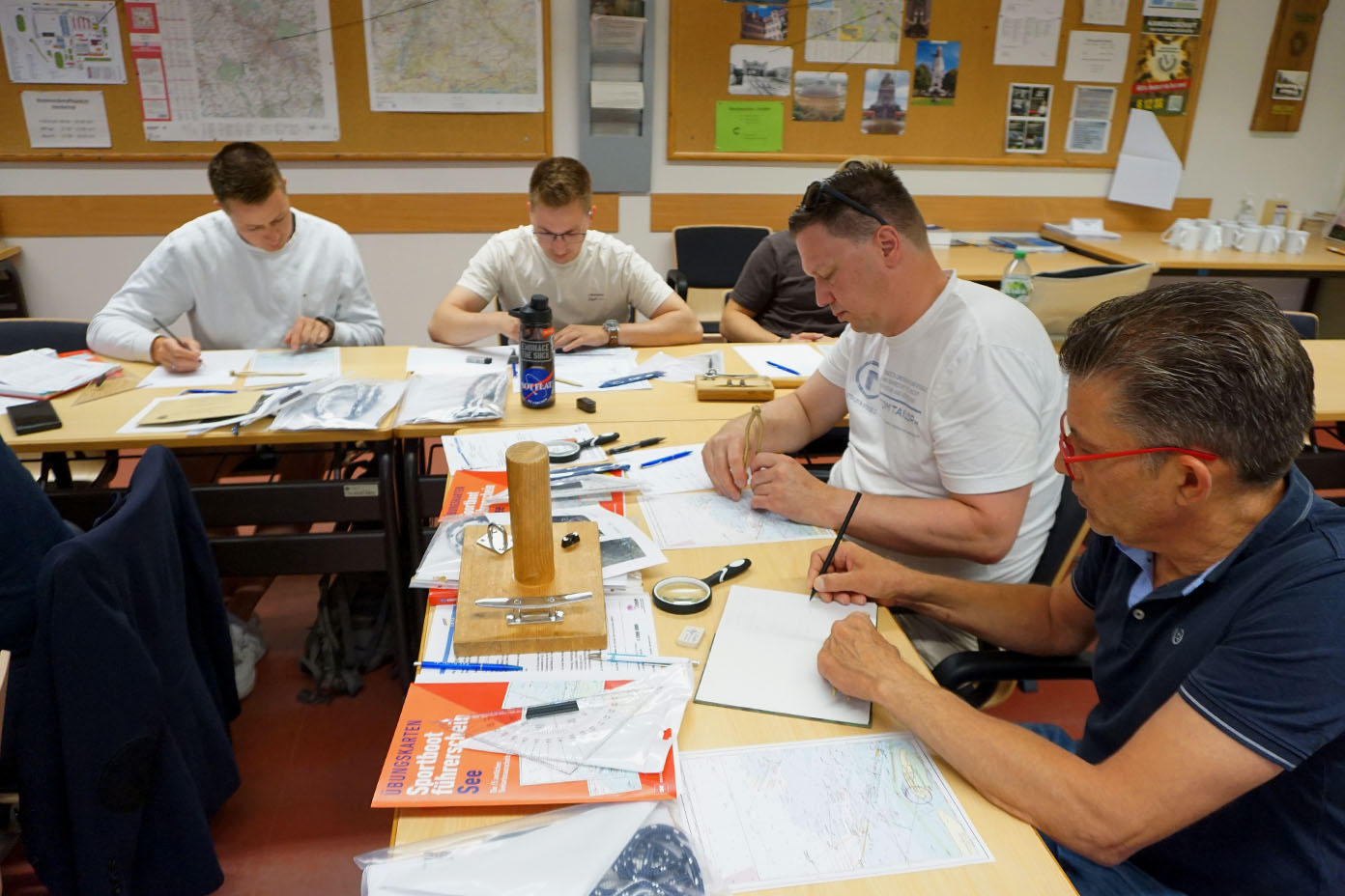 Vier Erwachsene sitzen an einem Tisch in einem Klassenraum und arbeiten konzentriert mit Stiften und Dreiecken auf Seekarten, umgeben von Karten, Büchern und Schreibmaterialien.