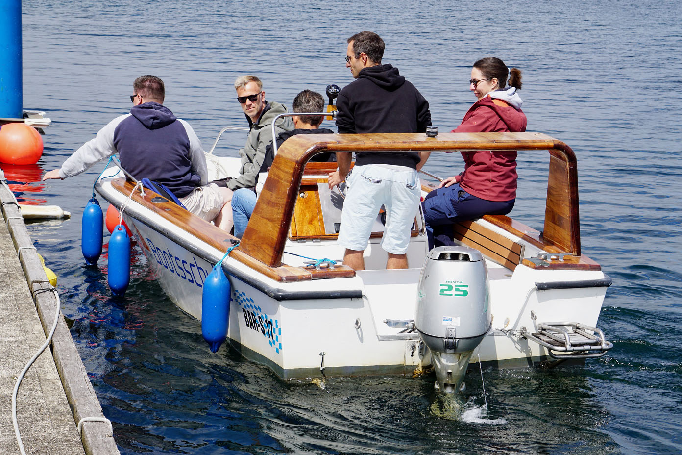 Fünf Personen sitzen und stehen auf einem kleinen Motorboot, das auf einem See an einem Steg anlegt.