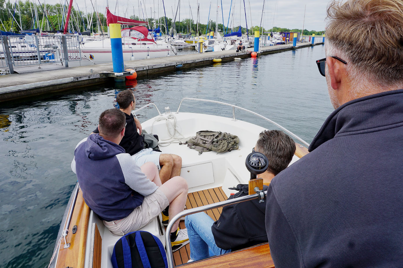 Vier Personen sitzen auf einem Boot im Hafen, umgeben von Wasser und anderen festgemachten Segelbooten.
