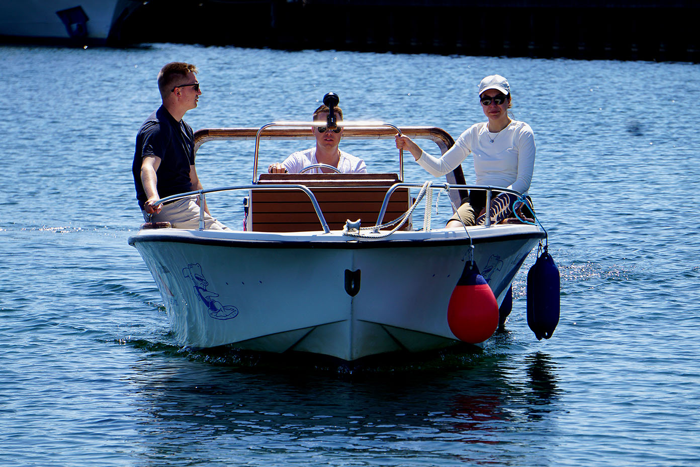 Drei Personen sitzen auf einem kleinen Boot auf ruhigem Wasser, eine trägt eine weiße Mütze und ein weißes Oberteil, während eine andere Person ein dunkles Oberteil trägt.