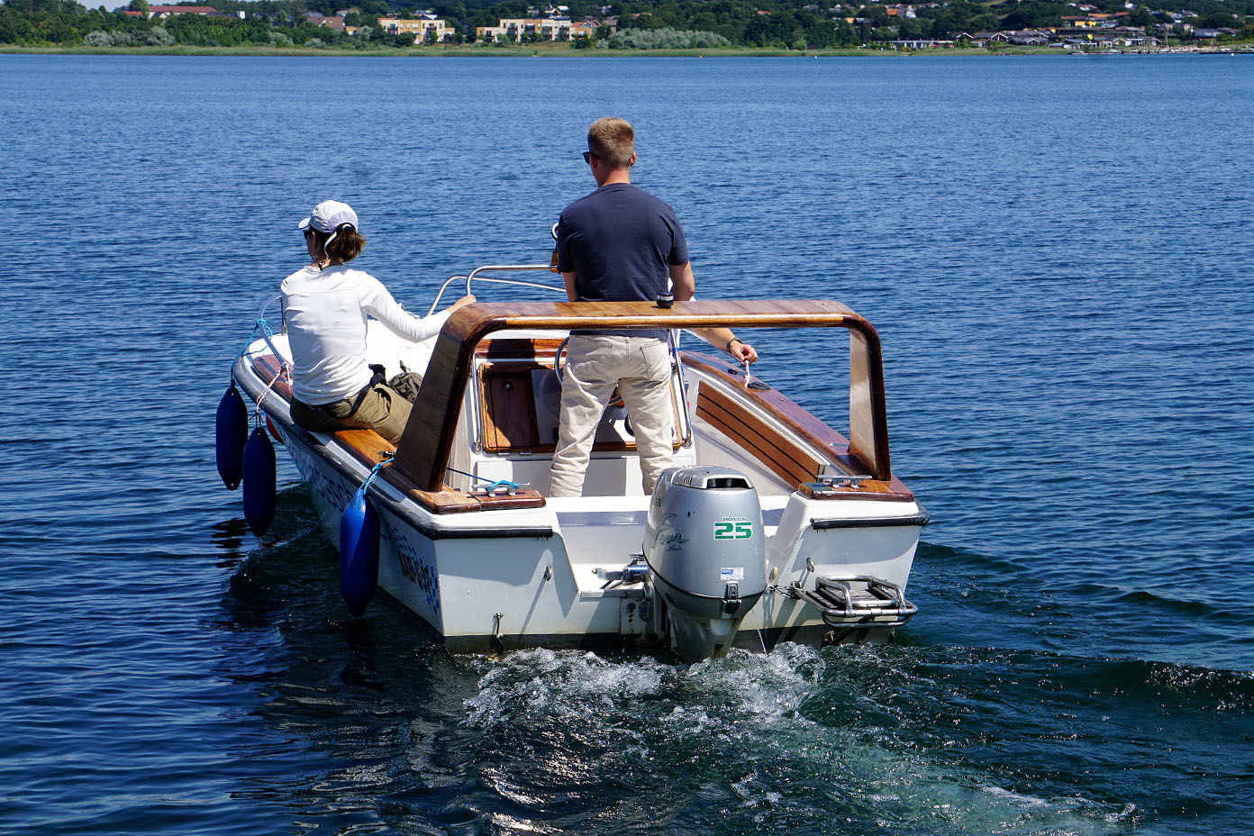 Zwei Personen sitzen auf einem kleinen Motorboot, das auf einem ruhigen See fährt, während eine Person steht und die Steuerung übernimmt.