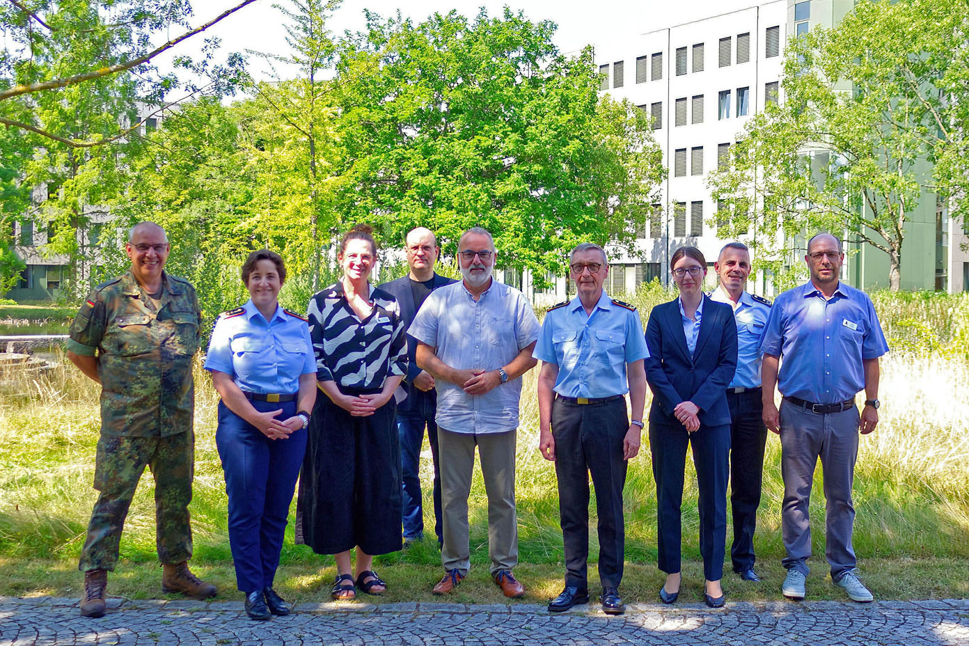 Neun Personen stehen in einer Reihe auf einem gepflasterten Weg vor einem Gebäude und grünen Bäumen, einige tragen formelle Kleidung und andere militärische Uniform.