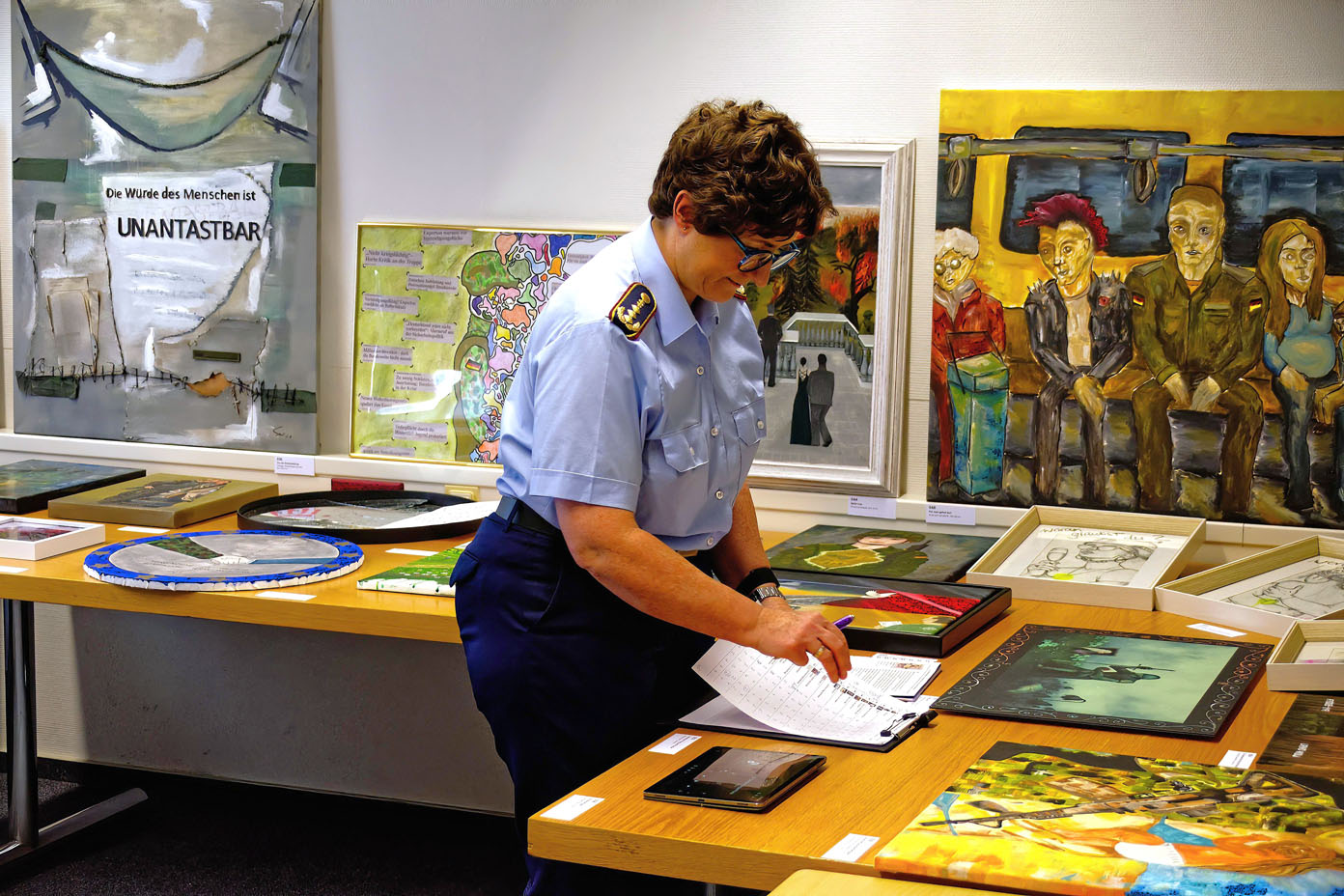Eine weibliche Person in militärischer Uniform steht an einem Tisch mit verschiedenen Kunstwerken und schreibt in ein Notizbuch.