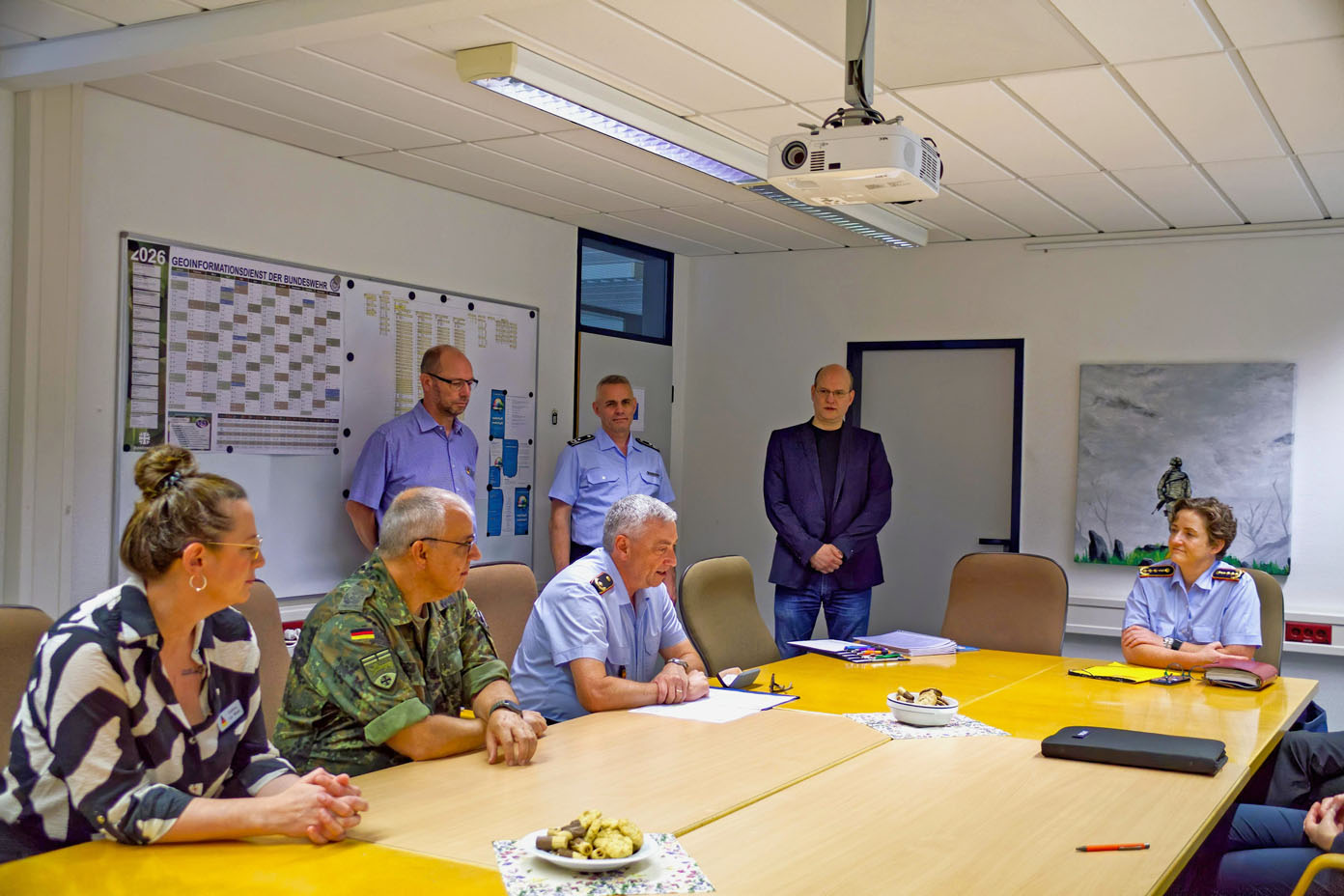 Mehrere Personen in Uniform und formeller Kleidung sitzen und stehen in einem Besprechungsraum um einen Tisch mit Unterlagen und kleinen Snacks.