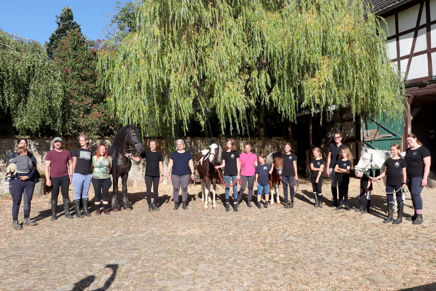 Eine Gruppe von Menschen steht nebeneinander auf einem gepflasterten Hof vor einem großen Baum und mehreren Pferden, einige tragen Reitstiefel und Freizeitkleidung.