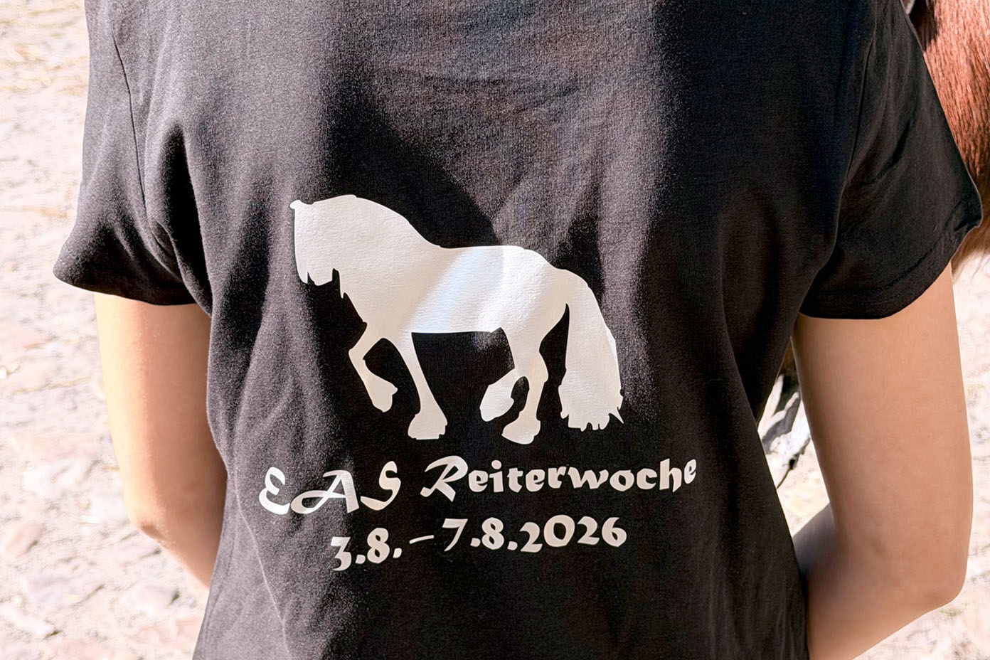 Eine Person trägt ein schwarzes T-Shirt mit einem weißen Pferde-Symbol und der Aufschrift EAS Reiterwoche 3.8. - 7.8.2026.
