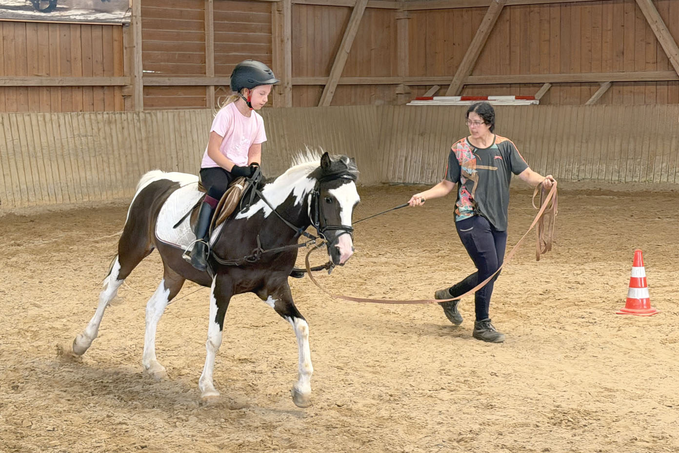 Ein Kind mit Helm reitet auf einem gescheckten Pferd in einer Reithalle, während eine Person das Pferd an einer langen Leine führt.