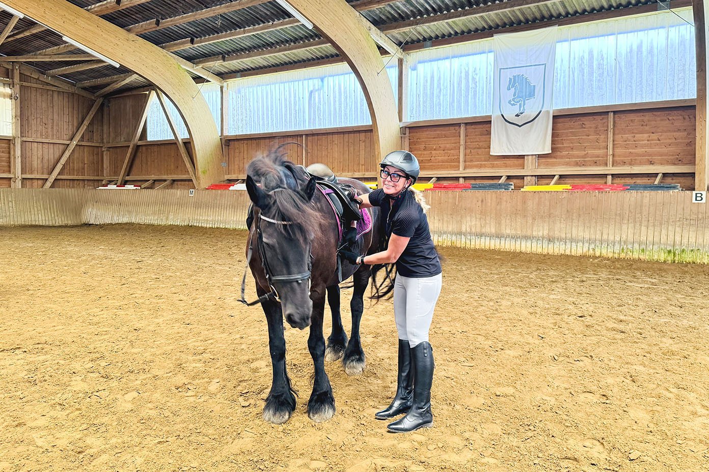 Eine Person in Reitkleidung und Helm steht in einer Reithalle neben einem gesattelten schwarzen Pferd und hält dessen Zügel.