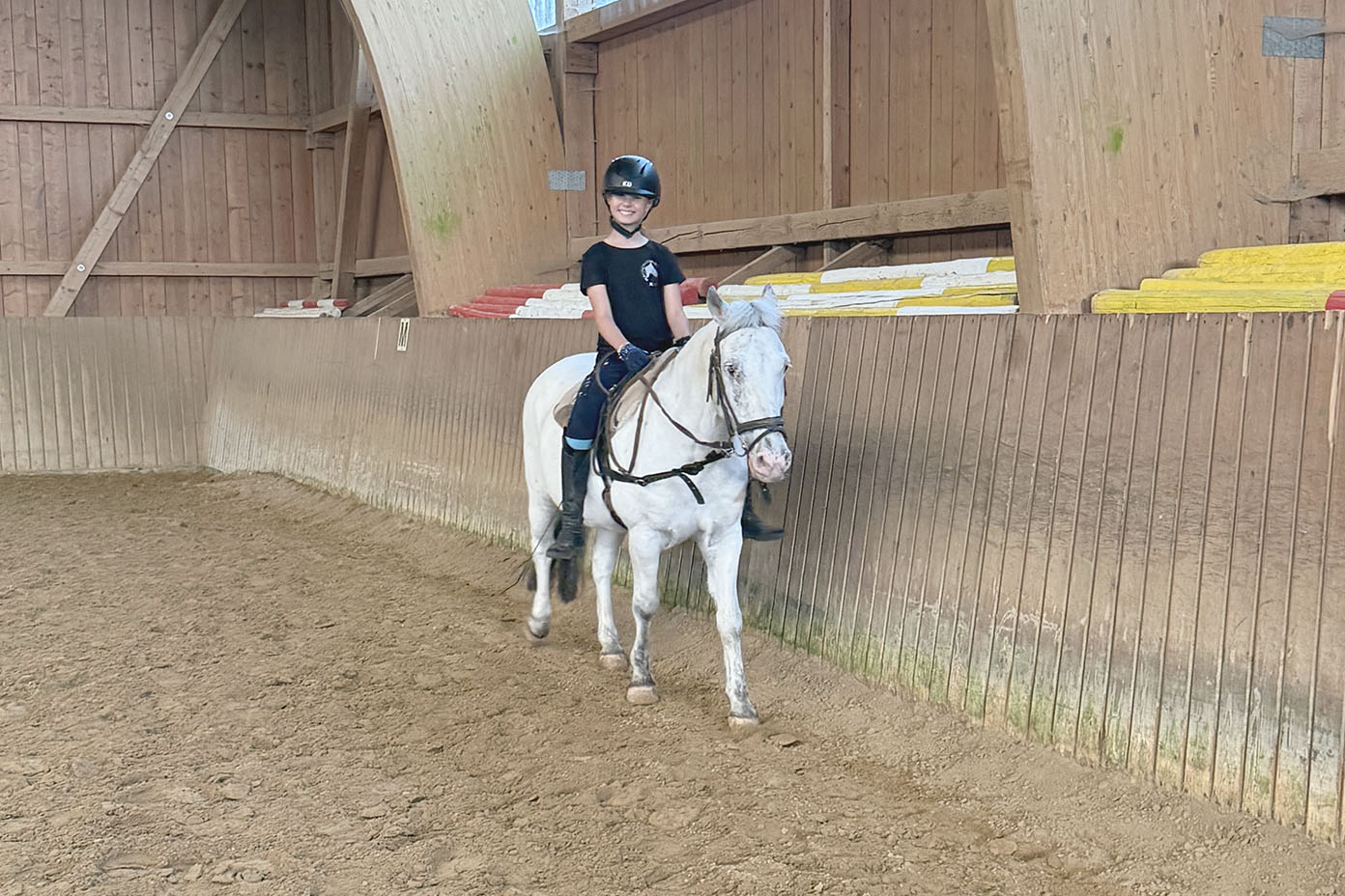 Eine Person mit Helm reitet in einer Reithalle auf einem weißen Pferd entlang einer Holzbarriere.