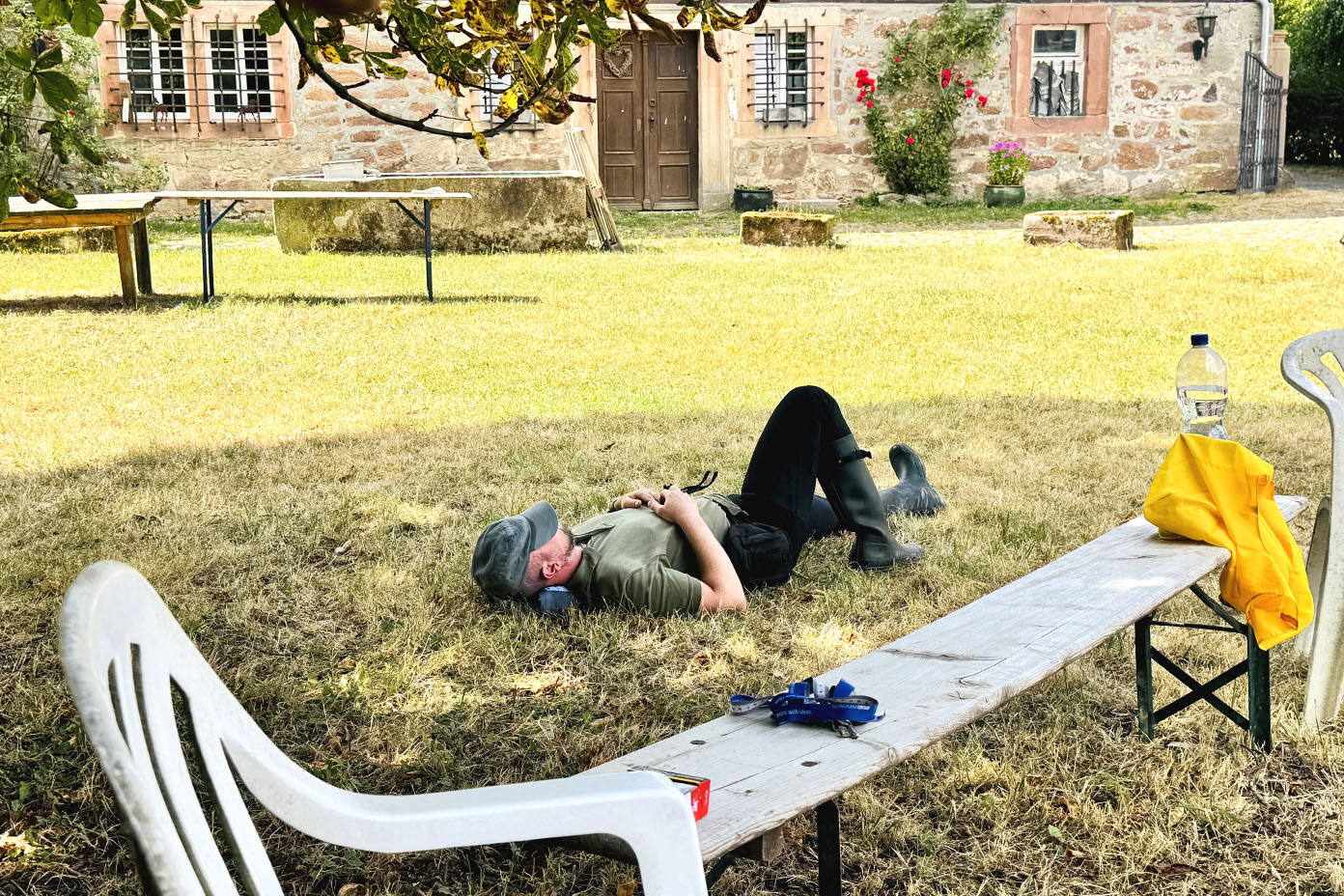 Eine Person liegt entspannt auf dem Rücken auf einer Wiese vor einem alten Steinhaus, umgeben von Gartenmöbeln und einer Wasserflasche.