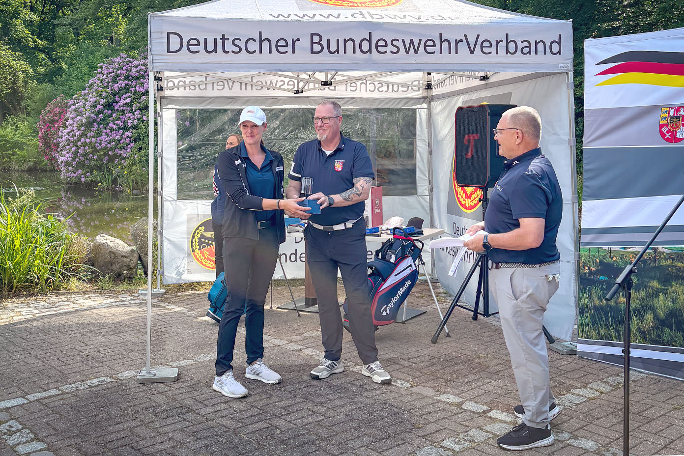 Drei Erwachsene stehen vor einem Zelt mit der Aufschrift Deutscher BundeswehrVerband, wobei zwei von ihnen sich die Hand geben und eine Trophäe übergeben wird.